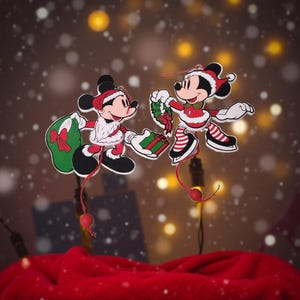 May include: Two Mickey Mouse Christmas ornaments. One Mickey holds a green bag with a red bow, the other holds a wreath and a gift. Both are wearing Santa hats and red and white outfits. The background is blurred with bokeh lights and falling snow.
