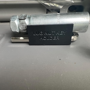 Pode incluir: Um suporte de plástico preto com o texto "LUG NUT KEY HOLDER" preso a uma chave de porca de roda prateada.