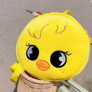 May include: A bright yellow plush backpack shaped like a cartoon chick. The backpack features large black eyes with white highlights, an orange beak, and small freckles. The bag has a yellow strap and is sitting in a woven basket.