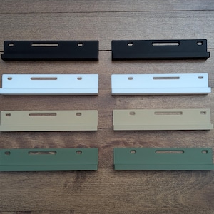 Puede incluir: Ocho escuadras de estantería rectangulares en negro, blanco, beige y verde. Cada escuadra tiene una ranura horizontal y dos orificios para el montaje. Las escuadras están dispuestas en pares sobre una superficie de madera.