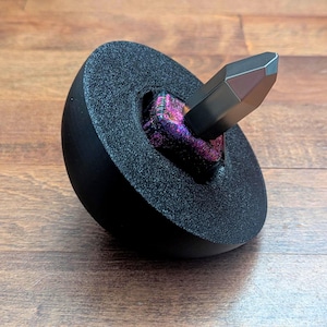 Pode incluir: Uma peão preto com uma parte superior pontiaguda metálica e iridescente. A peão está colocada numa base circular preta.