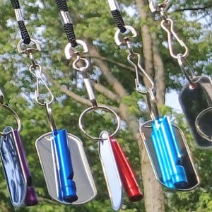 Op de afbeelding: Een verzameling zwarte lanyards met metalen clips en diverse gekleurde fluitjes. De lanyards hangen tegen een achtergrond van groen gebladerte, met fluitjes in blauwe, rode en paarse tinten.