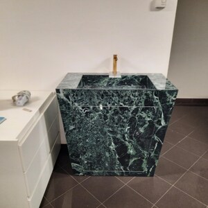 May include: A dark green marble sink with a gold faucet. The sink has a square basin and a trapezoidal base. The marble has a natural veining pattern. The sink is a modern bathroom fixture.