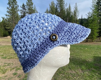May include: A blue and white crocheted hat with a button on the side. The hat has a wide brim and is made with a textured stitch.