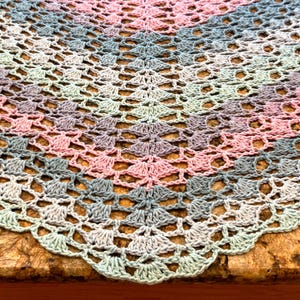 May include: A triangular crochet shawl featuring a repeating openwork pattern in a gradient of soft pastel colors including light pink, lavender, dusty blue, and pale mint green, finished with a scalloped edge along the bottom border.