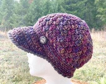May include: A crocheted hat with a wide brim and a button on the side. The hat is made in a variety of shades of purple, pink, and brown.