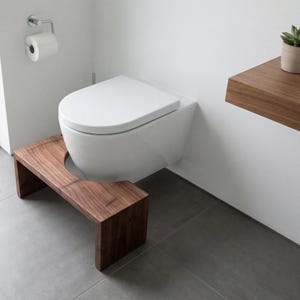 Könnte beinhalten: Eine weiße Wand-Toilette mit passendem weißem Sitz und Deckel. Direkt vor der Toilette steht ein dunkelbrauner Holzhocker mit einer geschwungenen Aussparung, die so geformt ist, dass sie um den Sockel der Toilette passt.