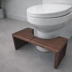 May include: A wooden toilet stool with a dark brown finish. The stool is rectangular with a curved cutout in the center to fit around the toilet base. It is designed to provide a comfortable and ergonomic boost.