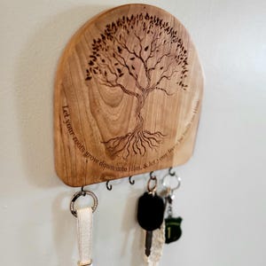 Op de afbeelding: Houten sleutelhouder met een boomontwerp en de tekst "Let your roots grow down into Him, & let your lives be built on Him." Inclusief meerdere haken voor sleutels. De sleutelhouder is gemaakt van lichtbruin hout.