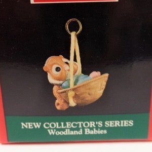 May include: A Hallmark Keepsake Ornament featuring a miniature woodland baby squirrel swinging in a basket. The box is red with the words "Hallmark Keepsake Ornament" and "New Collector's Series Woodland Babies" printed on it.