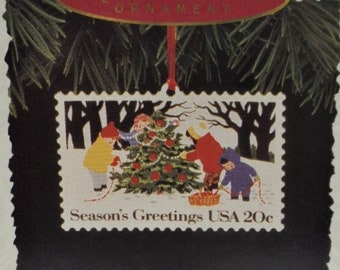 May include: A Keepsake Ornament featuring a vintage Christmas stamp with a scene of children decorating a Christmas tree. The stamp reads "Season's Greetings USA 20c". The ornament is part of the "U.S. Christmas Stamps" collector's series.