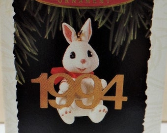 May include: White porcelain bunny ornament holding a gold 1994 charm. The ornament is in a white box with red trim and the words "Keepsake Ornament, Fabulous Decade, Collector's Series".