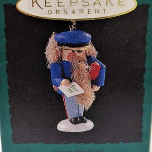May include: A Keepsake Ornament of a nutcracker soldier in a blue uniform, holding a red package and a small scroll that reads "Santa 1995". The ornament is in a green box with the text "Keepsake Ornament" and "Collector's Series".