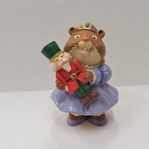 May include: A small, brown, ceramic figurine of a mouse wearing a purple dress and a crown, holding a nutcracker doll.