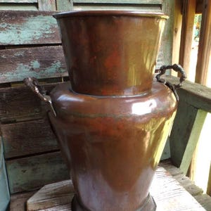 May include: A large, antique copper planter with a flared base and two ornate handles. The planter has a two-tiered design, with a smaller, bucket-shaped top section. The copper has a rich, aged patina, suggesting it is vintage.
