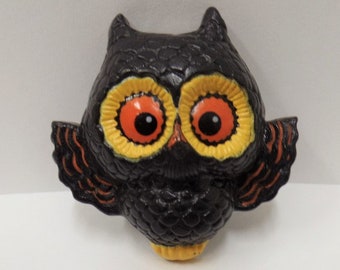 May include: A black ceramic owl figurine with orange and yellow painted eyes. The owl has black and orange wings and a yellow beak.