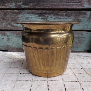 May include: A gold metal planter with a fluted design. The planter is sitting on a white crocheted doily.