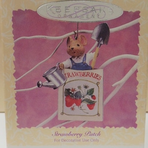 May include: A brown mouse wearing overalls holds a watering can and a shovel. The mouse is hanging from a branch with a sign that says "Strawberries" and has a picture of a strawberry plant. The sign is on a white background with a pink border. The text "Strawberry Patch" and "For Decorative Use Only" is printed below the image.