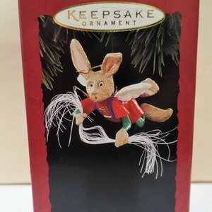 May include: A red and white box with the words "Keepsake Ornament" on the front. The box contains a Christmas ornament of a brown bunny with white wings and a halo. The bunny is holding a white string in its paws. The text "Angel Hare" is printed on the bottom of the box.