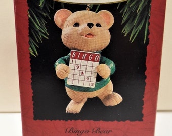 May include: A red box with a white border contains a brown bear holding a green bingo card. The card has the numbers 1, 9, 9, and 5. The box has the words "Keepsake Ornament" and "Bingo Bear" printed on it.