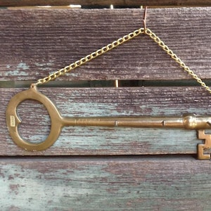 May include: A large brass key with a decorative design on the bottom. The key is hanging from a chain on a wooden surface.