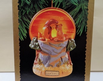 Puede incluir: Un "Keepsake Ornament" con Simba, Sarabi y Mufasa de "El Rey León". El adorno muestra a los personajes en una escena cálida iluminada por el sol. La base es una formación rocosa detallada. La caja es dorada con borde negro y detalles verdes.