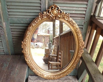 May include: A large, oval-shaped, gold-painted mirror with an ornate, floral-inspired frame. The mirror is hanging on a wooden wall with a slatted design.