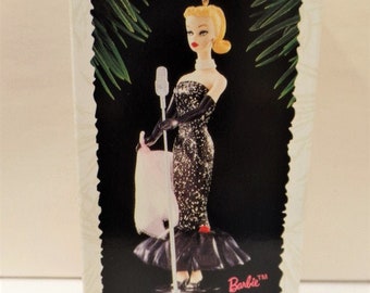 May include: A vintage Barbie doll Christmas ornament in a black sequined dress and black gloves, holding a microphone. The ornament is in its original box, which reads "Keepsake Ornament" and "Solo in the Spotlight" with the Barbie logo. The box is red, white, and black.