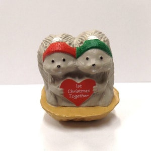 May include: A ceramic figurine of two gray hedgehogs wearing red and green hats, holding a red heart with the text "1st Christmas Together". The hedgehogs are sitting on a brown, nut-shaped base.