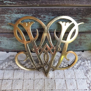 Puede incluir: Un salvamanteles de metal dorado con un diseño decorativo que presenta las letras "W" y "F" entrelazadas. El salvamanteles tiene forma de corazón y un diseño delicado y ornamentado.