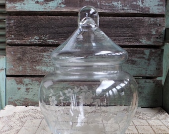 May include: A clear glass apothecary jar with a lid. The jar has a rounded body and a lid with a small handle. The glass is etched with a floral design. The jar is sitting on a cream-colored, patterned tablecloth.