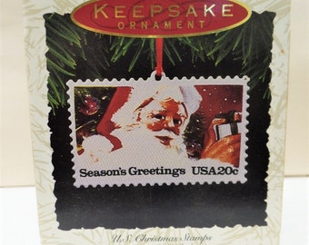 May include: A red and white Christmas ornament featuring a vintage US postage stamp with Santa Claus and the text "Season's Greetings USA 20c". The ornament is part of a "New Collector's Series" of US Christmas stamps.