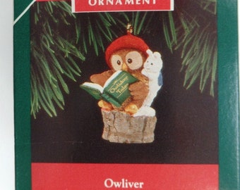 May include: A Hallmark Keepsake ornament featuring an owl wearing a red hat, reading a book titled "Holiday Tales." The ornament includes a white bunny and is set on a brown base. The box reads "New Collector's Series" and "Owlver."