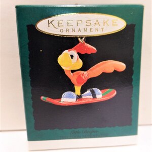 May include: A red and yellow cartoon rooster wearing blue and black boots is riding a red and green snowboard. The ornament is in a green box with the words "Keepsake Ornament" on it. The box also has the words "Little & Brother" on it.