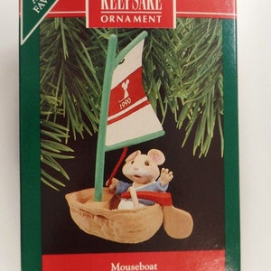 May include: A Hallmark Artists' Favorites Keepsake Ornament featuring a mouse in a sailboat. The ornament is red, white, and blue with the year 1990 on the sail. The box reads "Mouseboat Sculpted by Ed Seale".