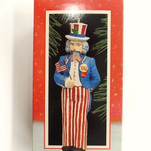 May include: A vintage Uncle Sam Nutcracker ornament in red, white, and blue. The ornament is wearing a top hat with a red, white, and blue band and a green holly sprig. The ornament is holding a small American flag and has a blue jacket with a red, white, and blue striped shirt and pants. The ornament has a white beard and mustache. The box reads "Uncle Sam Nutcracker Handcrafted Ornament Dated 1988".