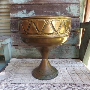 May include: A gold-toned metal planter with a decorative geometric pattern around the rim. The planter has a wide, round base and a tall, slender stem.
