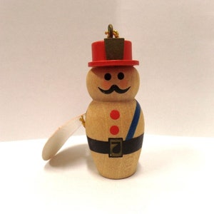 May include: Wooden figurine of a person wearing a red hat, black belt, and blue sash. The figurine has a painted face with a mustache and two red dots.