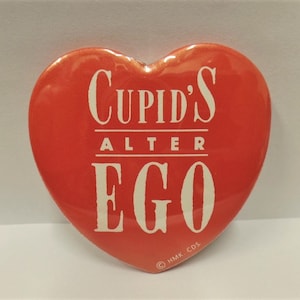 May include: Red heart-shaped button with white text that reads "Cupid's Alter Ego" and a copyright symbol with "HMK. CDS."
