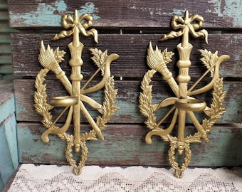 May include: A pair of gold-toned metal wall sconces with a decorative design featuring a bow, arrows, and a brush. Each sconce has a small circular candle holder.