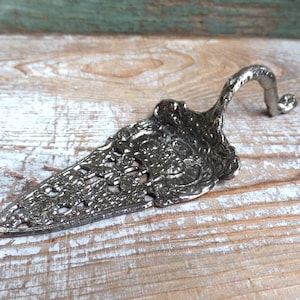 May include: Silver-toned cake server with an ornate, openwork design on the blade. The handle curves upward and ends in a decorative flourish. The server is made of metal and rests on a wooden surface.