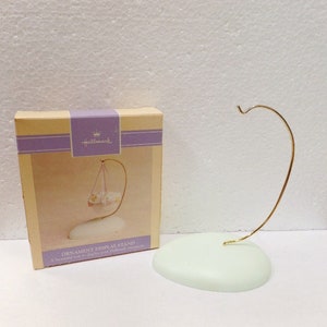 May include: A white ceramic ornament display stand with a gold metal hook. The stand is shaped like a rounded oval and has the text "ORNAMENT DISPLAY STAND" and "A beautiful way to display your Hallmark ornaments" printed on the box.