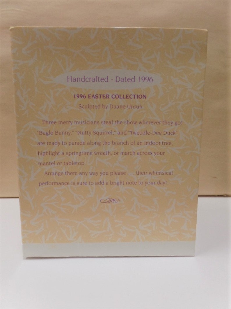 Puede incluir: Una tarjeta con fondo amarillo claro y texto blanco. El texto dice "Handcrafted - Dated 1996", "1996 Easter Collection", "Sculpted by Duane Unruh", "Three merry musicians steal the show wherever they go!" "Bugle Bunny", "Nutty Squirrel", and "Tweedle-Dee Duck" are ready to parade along the branch of an indoor tree, highlight a springtime wreath, or march across your mantel or tabletop. Arrange them any way you please - their whimsical performance is sure to add a bright note to your day!"