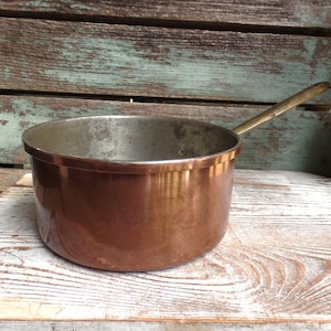 May include: A copper saucepan with a brass handle. The saucepan is sitting on a wooden surface.