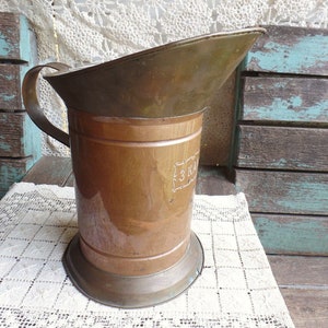 May include: A copper pitcher with a wide spout and a handle. The pitcher has a copper base and a copper body with a band of copper around the middle. The pitcher has a label on the side that says "3 KILO".