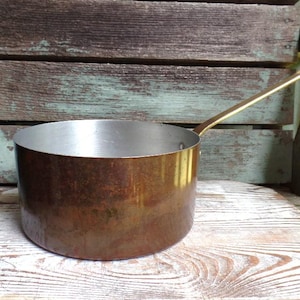 May include: A vintage copper saucepan with a long brass handle. The pot has a rounded shape and a shiny interior. The exterior of the pot has a warm, aged patina. The handle extends to the right, suggesting it's ready for use.