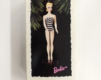 May include: A white box with a red border and the text "Keepsake Ornament" in black. The box features a black and white striped Barbie doll ornament with blonde hair and a black and white swimsuit. The text "Barbie" is in pink and the text "Debut - 1959" is in black. The text "New Collector's Series" is in white on a red background.