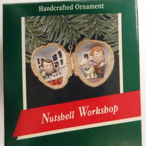 May include: A handcrafted ornament in the shape of a walnut shell with a hinged opening. The ornament features two miniature figures inside, one holding a small tree and the other holding a book. The ornament is labeled "Nutshell Workshop".