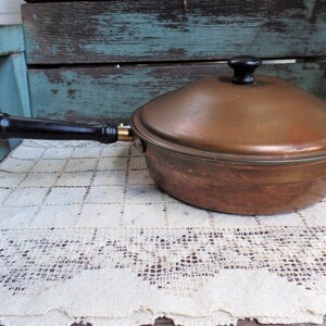 May include: A vintage copper saucepan with a lid and a black wooden handle. The saucepan is sitting on a white crocheted doily.