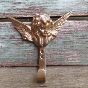 May include: A brass hook with an angel cherub design. The angel has wings and a face with a sweet expression. The hook is a decorative and functional piece.